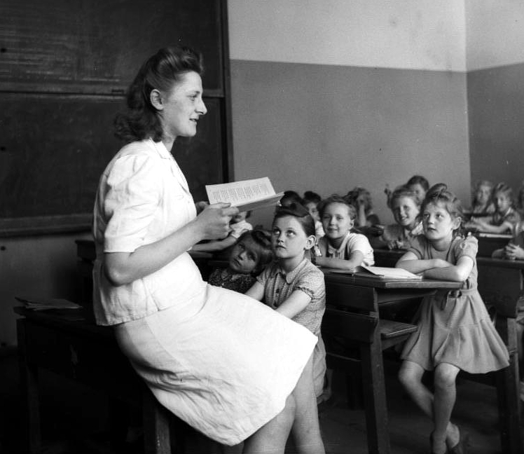 Eine Lehrerin vor einer Klasse Grundschulkinder. Die Kinder schauen sie an, sie schaut mit geöffnetem Buch über sie hinweg.